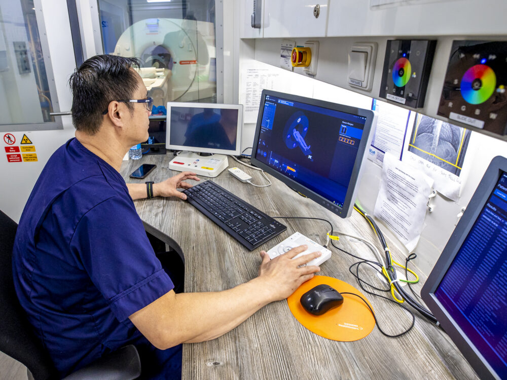 NHS healthcare professional reviewing medical scan images on a computer in a hospital imaging control room.